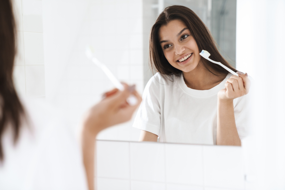 young woman smiling and brushing her teeth in mirror